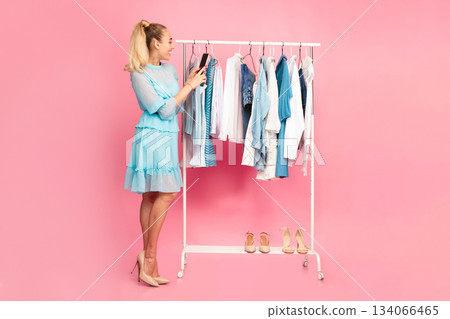 Vertical full length portrait of smiling young woman holding mobile phone and taking photo picture of clothes, recording video blog standing near clothing rail, selling used shoes and dresses 134066465