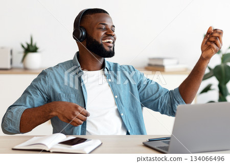 Work With Fun. Cheerful Black Freelancer Man In Wireless Headphones Listening Music And Playing Virtual Guitar At Workplace, Resting At Desk In Home Office, Enjoying Remote Job Opportunities 134066496