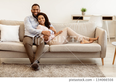 Beautiful african american black man and woman relaxing on couch at home in living room, smiling couple looking at camera and posing, spending leisure time together, watching TV or movie, embracing 134066508