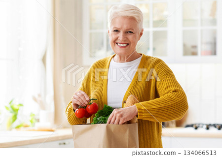 Happy senior woman checking paper bag with grocery ordered from internet during COVID-19 pandemic, kitchen interior, copy space. Smiling old lady unpacking purchase after shopping. Food delivery 134066513