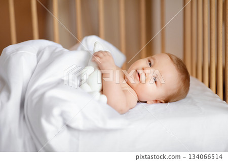 Good morning world. Happy baby boy waking up after sleeping in wooden crib, laying under blanket and smiling, holding white toy, side view. Cute baby girl laying in toddler bed 134066514