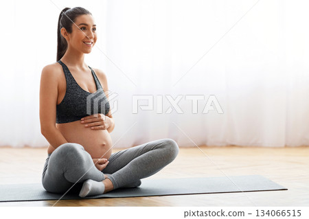 Young expecting woman training on mat at home, getting ready for childbirth. Smiling beautiful pregnant female exercising in living room, tenderly embracing tummy and looking away, copy space Young expecting woman training on mat at home, getting ready for childbirth. Smiling beautiful pregnant female exercising in living room, tenderly embracing tummy and looking away, copy space 134066515