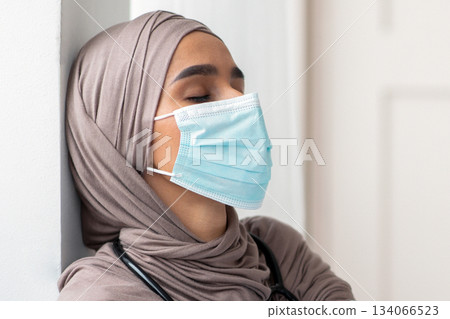 Muslim woman doctor feeling exhausted, wearing protective face mask and gloves, working hard during pandemic. Physician lady in hijab sitting on floor by wall in her cabinet, copy space Muslim woman doctor feeling exhausted, wearing protective face mask and gloves, working hard during pandemic. Physician lady in hijab sitting on floor by wall in her cabinet, copy space 134066523