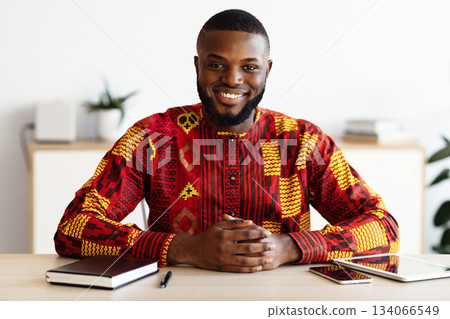 Portrait Of Handsome African Freelancer Man In Traditional Costume Sitting At Desk In Home Office, Positive Black Self-Employed Guy In Ethnic Costume Smiling And Looking At Camera, Free Space 134066549