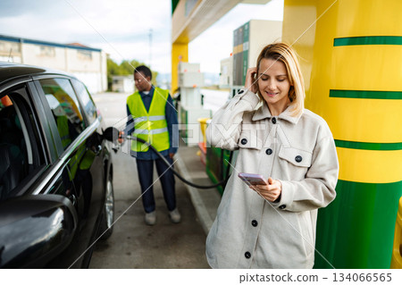 Woman using smartphone while gas station attendant refueling car Woman using smartphone while gas station attendant refueling car 134066565