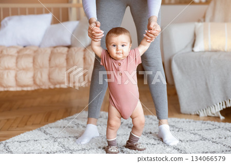 Cute little baby boy or girl taking first steps with mother help, home interior, close up. Adorable toddler holding mom hands, little blonde kid daughter or son walking by living room Cute little baby boy or girl taking first steps with mother help, home interior, close up. Adorable toddler holding mom hands, little blonde kid daughter or son walking by living room 134066579