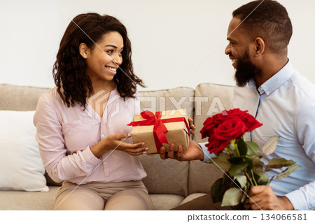 Celebrating Special Occasion. Portrait of smiling african american guy giving his girlfriend wrapped box with present and a bouquet of red roses. Happy man greeting lady with international women's day Celebrating Special Occasion. Portrait of smiling african american guy giving his girlfriend wrapped box with present and a bouquet of red roses. Happy man greeting lady with international women's day 134066581