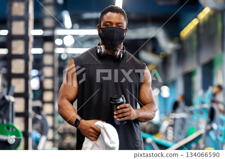 Active lifestyle during pandemic. African american muscular man with wireless headphones wearing protective mask while exercising at gym, holding protein drink and towel, break time, copy space Active lifestyle during pandemic. African american muscular man with wireless headphones wearing protective mask while exercising at gym, holding protein drink and towel, break time, copy space 134066590