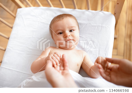Good morning sunshine. Beautiful little baby laying in cot, holding mom's hands, ready to wake up. Top view of cute blonde toddler waking up, lying in wooden baby bed, grabbing mother fingers Good morning sunshine. Beautiful little baby laying in cot, holding mom's hands, ready to wake up. Top view of cute blonde toddler waking up, lying in wooden baby bed, grabbing mother fingers 134066598
