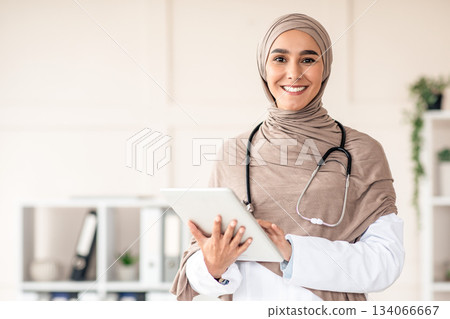 Modern technologies in healthcare concept. Happy female doctor in hijab with digital tablet posing in her office, copy space. Muslim yougn woman therapist holding digital pad, clinic interior Modern technologies in healthcare concept. Happy female doctor in hijab with digital tablet posing in her office, copy space. Muslim yougn woman therapist holding digital pad, clinic interior 134066667