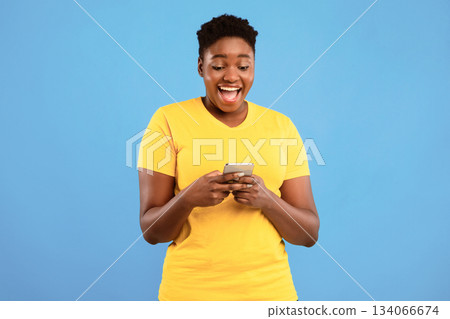 Excited African Plus-Size Woman Using Phone Application Standing Over Blue Studio Background. Black Lady Playing Mobile Game On Cellphone. Great App For Your Smartphone Concept Excited African Plus-Size Woman Using Phone Application Standing Over Blue Studio Background. Black Lady Playing Mobile Game On Cellphone. Great App For Your Smartphone Concept 134066674