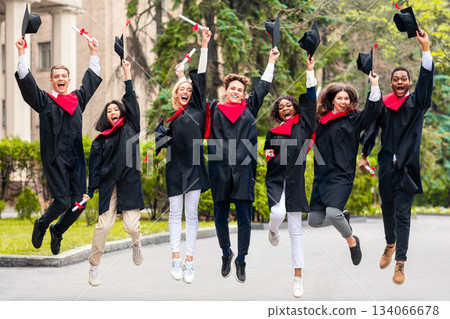 Happy international students celebrating graduation from university or college, holding diplomas and graduation hats, jumping up, laughing and posing at camera, campus background 134066678
