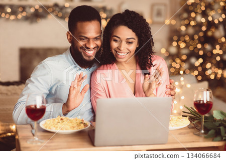 Online Celebration And Communication. Smiling african american man and woman sitting at the table during online video chat with friends or family using laptop, waving to webcam, having dinner 134066684