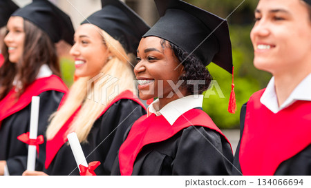 International group of students having graduation ceremony, wearing graduation costumes and holding diplomas, standing in a row and cheerfully smiling, closeup, panorama 134066694