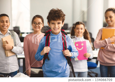 Return Back To School. Smiling small boy wearing backpack posing in classroom, diverse group of happy international classmates standing in line in the blurred background, studying and learning Return Back To School. Smiling small boy wearing backpack posing in classroom, diverse group of happy international classmates standing in line in the blurred background, studying and learning 134066709