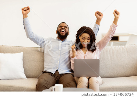 Yeah. Portrait of excited black couple celebrating online win, great deal or business success, using laptop computer, screaming with clenched fists sitting on the sofa in living room at home Yeah. Portrait of excited black couple celebrating online win, great deal or business success, using laptop computer, screaming with clenched fists sitting on the sofa in living room at home 134066724