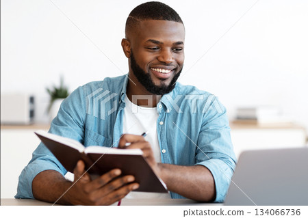 Black Millennial Freelancer Guy Taking Notes To Notepad And Working On Laptop At Home Office, Young African American Man Looking At Computer Screen And Writing In Notepad, Planning Business Schedule 134066736