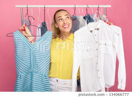 Dilemma Concept. Portrait of young pensive lady choosing trendy outfit, holding hangers with dress and shirt, thinking about purchase, making decision what to wear, rack in the background 134066777
