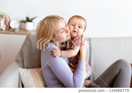 Happy motherhood. Affectionate young mother blonde woman cuddling with her cute little baby, sitting on sofa in living room. Beautiful mom and happy sweet kid toddler hugging at home 134066779