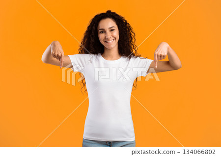Happy Curly Woman Pointing Fingers Down Posing Smiling To Camera Standing In Studio Over Yellow Background, Wearing White T-Shirt. Look There Advertisement Banner. Panorama Happy Curly Woman Pointing Fingers Down Posing Smiling To Camera Standing In Studio Over Yellow Background, Wearing White T-Shirt. Look There Advertisement Banner. Panorama 134066802