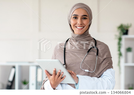 Modern technologies in healthcare concept. Happy female doctor in hijab with digital tablet posing in her office, copy space. Muslim yougn woman therapist holding digital pad, clinic interior Modern technologies in healthcare concept. Happy female doctor in hijab with digital tablet posing in her office, copy space. Muslim yougn woman therapist holding digital pad, clinic interior 134066804