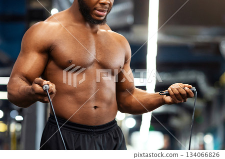 Hot african american young man bodybuilder warming up before working out in gym, closeup. Shirtless muscular black guy jumping with skipping, looking at free space while making cardio Hot african american young man bodybuilder warming up before working out in gym, closeup. Shirtless muscular black guy jumping with skipping, looking at free space while making cardio 134066826
