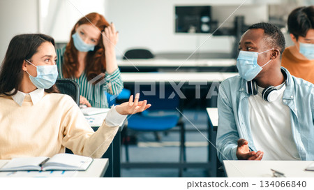 Education, Lifestyle And People Concept. Portrait of smiling diverse international students wearing protective medical masks and talking, sitting at desks in lecture hall at the university, discussing 134066840