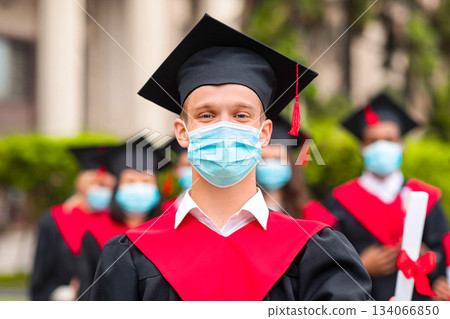 COVID-19 pandemic and education. Guy student in graduation costume showing diploma at camera, posing over international group of students at university campus, wearing protective face mask, copy space 134066850