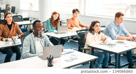 Lecture Concept. Group of interested multicultural mixed race group of students sitting at tables with pc in modern classroom, listening to teacher, taking notes writing in notebooks, back to study Lecture Concept. Group of interested multicultural mixed race group of students sitting at tables with pc in modern classroom, listening to teacher, taking notes writing in notebooks, back to study 134066860