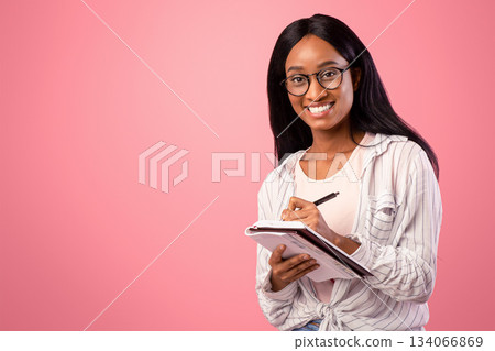 Portrait of intelligent black female student wearing glasses, taking notes in copybook on pink studio background. Smart African American woman studying, preparing for exam, writing in notebook Portrait of intelligent black female student wearing glasses, taking notes in copybook on pink studio background. Smart African American woman studying, preparing for exam, writing in notebook 134066869