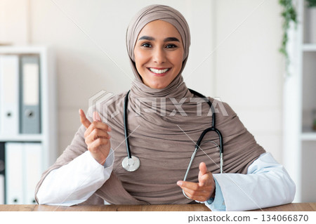 Smiling female doctor young beautiful woman in hijab with stethoscope making consultation in her cabinet, sitting at workdesk with medical chart, notebook and laptop in clinic, copy space Smiling female doctor young beautiful woman in hijab with stethoscope making consultation in her cabinet, sitting at workdesk with medical chart, notebook and laptop in clinic, copy space 134066870