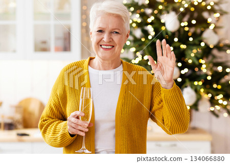 Positive mature lady in celebrating New Year or xmas with her family or friends online, having video call, waving at camera, holding glass of champagne, decorated kitchen interior, empty space 134066880