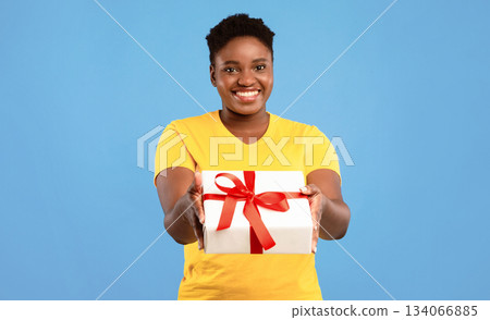 Cheerful Plus Size African Woman Offering Wrapped Gift Box To Camera Posing Standing Over Blue Studio Background. This Present Is For You. Birthday Celebration, Holiday Gifts And Presents Concept Cheerful Plus Size African Woman Offering Wrapped Gift Box To Camera Posing Standing Over Blue Studio Background. This Present Is For You. Birthday Celebration, Holiday Gifts And Presents Concept 134066885