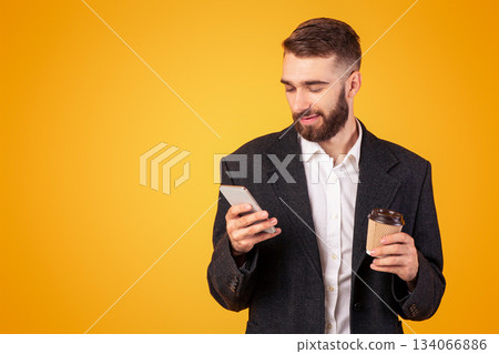 Positive young businessman checking email or browsing web on his cellphone, drinking takeaway coffee on orange studio background. Happy guy in formal wear using internet on mobile device 134066886