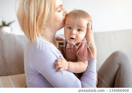 Beautiful lady bonding with her adorable baby, holding cute sun or daughter and kissing, sitting on couch. Closeup portrait of loving mother young blonde woman kissing little kid, home interior Beautiful lady bonding with her adorable baby, holding cute sun or daughter and kissing, sitting on couch. Closeup portrait of loving mother young blonde woman kissing little kid, home interior 134066908