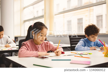 Learning Process Concept. Portrait of Small Schoolchildren Smiling, Sitting At Desk Taking Notes And Writing in Exercise Copybook. Junior Classroom with Diverse Group of Children Learning New Stuff Learning Process Concept. Portrait of Small Schoolchildren Smiling, Sitting At Desk Taking Notes And Writing in Exercise Copybook. Junior Classroom with Diverse Group of Children Learning New Stuff 134066912