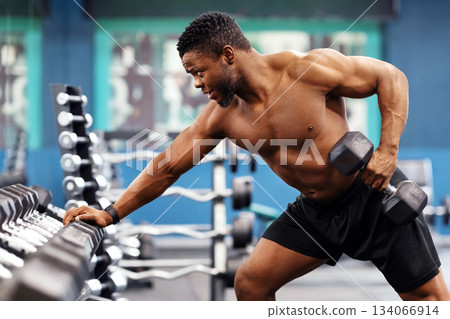Motivated african american shirtless bodybuilder training arms and back muscles with dumbbells at modern gym, empty space. Hot black young man working on his biceps, lifting barbell with one hand Motivated african american shirtless bodybuilder training arms and back muscles with dumbbells at modern gym, empty space. Hot black young man working on his biceps, lifting barbell with one hand 134066914