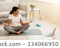 Yoga Online. Relaxed young african american woman meditating in front of laptop at home, sitting in lotus position on mat and doing breathing exercises in living room or bedroom, free space Yoga Online. Relaxed young african american woman meditating in front of laptop at home, sitting in lotus position on mat and doing breathing exercises in living room or bedroom, free space 134066950
