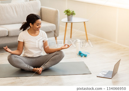 Yoga Online. Relaxed young african american woman meditating in front of laptop at home, sitting in lotus position on mat and doing breathing exercises in living room or bedroom, free space Yoga Online. Relaxed young african american woman meditating in front of laptop at home, sitting in lotus position on mat and doing breathing exercises in living room or bedroom, free space 134066950