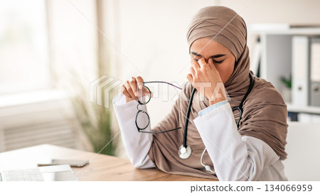 Muslim doctor young woman in hijab with stethoscope having eyesight problem while working with laptop at clinic, holding glasses and rubbing her nose bridge with closed eyes, copy space 134066959