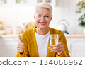 Cheerful senior grey-haired woman with short haircut holding glass of water showing thumb up, enjoying healthy lifestyle while retirement, sitting at white kitchen at home, copy space Cheerful senior grey-haired woman with short haircut holding glass of water showing thumb up, enjoying healthy lifestyle while retirement, sitting at white kitchen at home, copy space 134066962