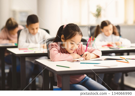 Education And Studying Concept. Portrait of smiling asian small student sitting at desk in classroom, writing in notebook or drawing picture with colorful pencils. Back To School 134066990