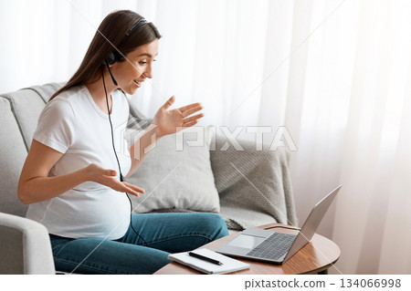 Remote Work. Happy Young Pregnant Female Tutor Making Online Video Lesson With Laptop, Wearing Headset, Talking And Gesturing At Computer Web Camera, Sitting On Couch In Living Room, Side View 134066998