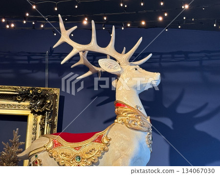White deer statue with ornate golden harness standing under festive string lights. Majestic holiday presence, elegant seasonal display, decorative interior charm and Christmas atmosphere mood White deer statue with ornate golden harness standing under festive string lights. Majestic holiday presence, elegant seasonal display, decorative interior charm and Christmas atmosphere mood 134067030