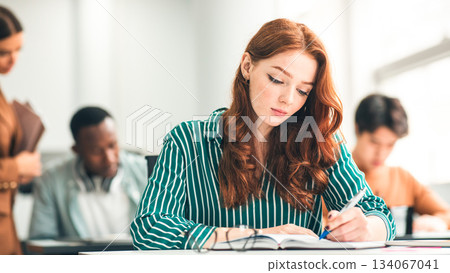 People And Studying Concept. Lifestyle portrait of pensive diligent redhead lady sitting at desk in academic auditorium hall, taking exam, thinking about question and task. Higher education 134067041