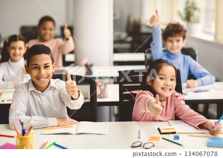 Education Is Fun Concept. Diverse group of happy smiling international classmates sitting at desks in classroom and showing thumbs up sign gesture, children enjoying studying at junior school 134067078