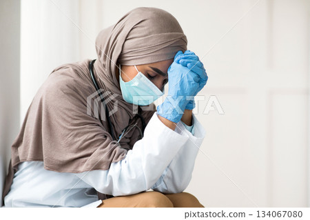 Sad woman in hijab doctor having difficult day at clinic, wearing protective gloves and face mask, sitting next to wall in her office and resting, empty space. Upset female doctor feeling exhausted Sad woman in hijab doctor having difficult day at clinic, wearing protective gloves and face mask, sitting next to wall in her office and resting, empty space. Upset female doctor feeling exhausted 134067080