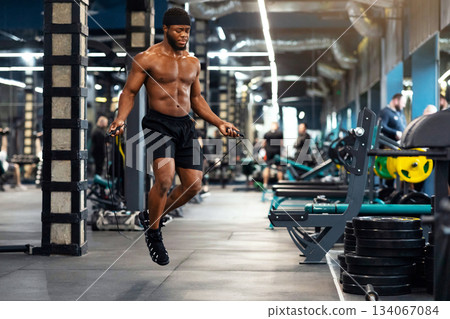 Cardio workout. Shirtless muscular african american man jumping with skipping rope in gym interior, looking at free space. Hot black guy bodybuilder warming up before working out Cardio workout. Shirtless muscular african american man jumping with skipping rope in gym interior, looking at free space. Hot black guy bodybuilder warming up before working out 134067084