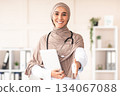 Modern technologies in healthcare concept. Happy female doctor in hijab with digital tablet greeting someone in her office, copy space. Muslim woman therapist holding digital pad, clinic interior Modern technologies in healthcare concept. Happy female doctor in hijab with digital tablet greeting someone in her office, copy space. Muslim woman therapist holding digital pad, clinic interior 134067088