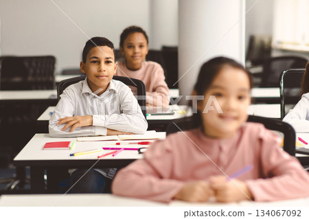 Elementary Education. Portrait of small smiling boy sitting at table in classroom at primary school or kindergarten with group of classmates. Return back to school after covid quarantine Elementary Education. Portrait of small smiling boy sitting at table in classroom at primary school or kindergarten with group of classmates. Return back to school after covid quarantine 134067092